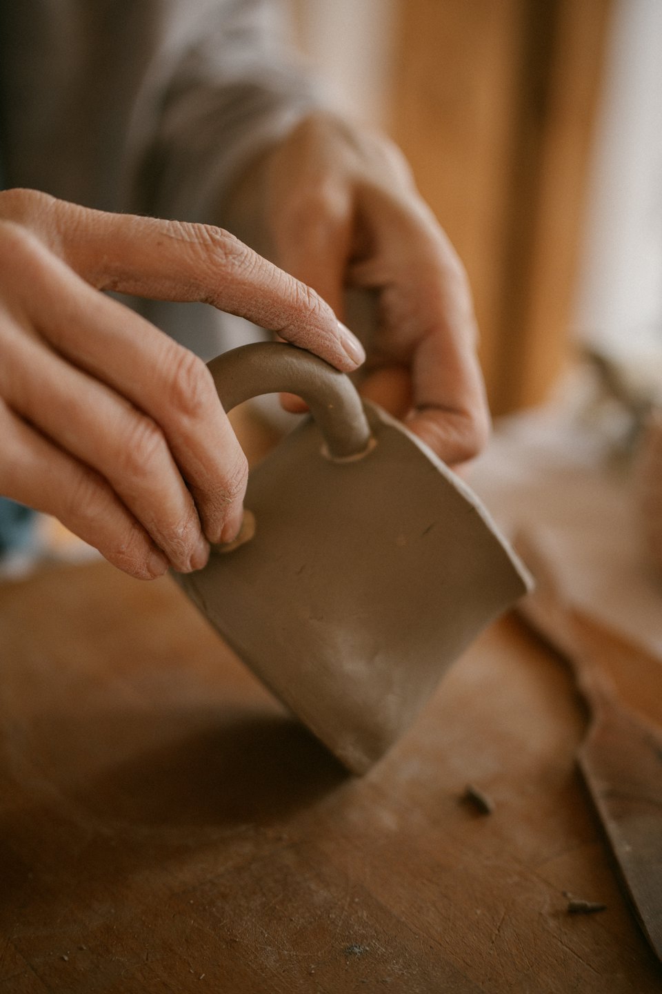 Hands shaping a clay mug handle on a wooden workbench.